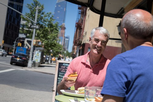 Two men sit at an outdoor café table, enjoying lunch and chatting, with a cityscape in the background.