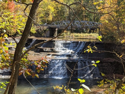 A serene waterfall cascades over rocks, framed by autumn foliage and a bridge in the background.