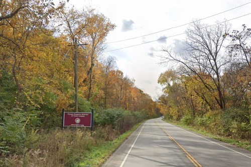 A scenic road lined with trees displaying autumn colors, with a sign for a nature preserve on the side.
