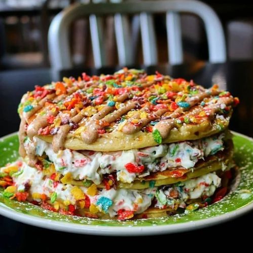 A colorful stack of pancakes layered with cream and topped with fruity cereal and drizzled sauce on a green plate.