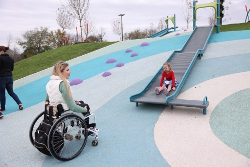 A woman in a wheelchair watches a child sitting on a slide in a colorful playground.