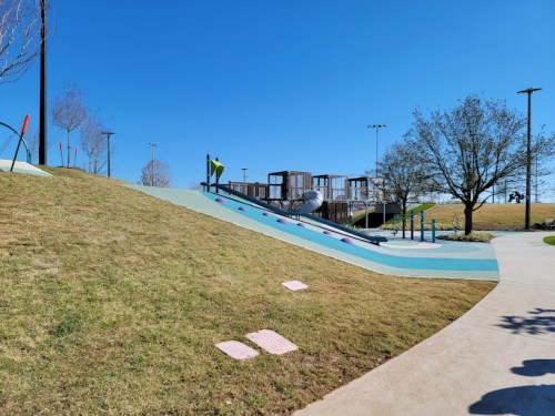 A sunny park scene featuring a grassy hill, a slide, and playground equipment in the background.
