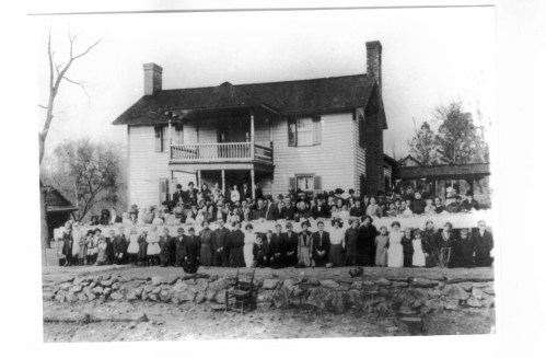 A large group of people, including men, women, and children, poses in front of a two-story house.