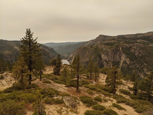 A scenic view of a mountainous landscape with pine trees and a river winding through the valley under a hazy sky.