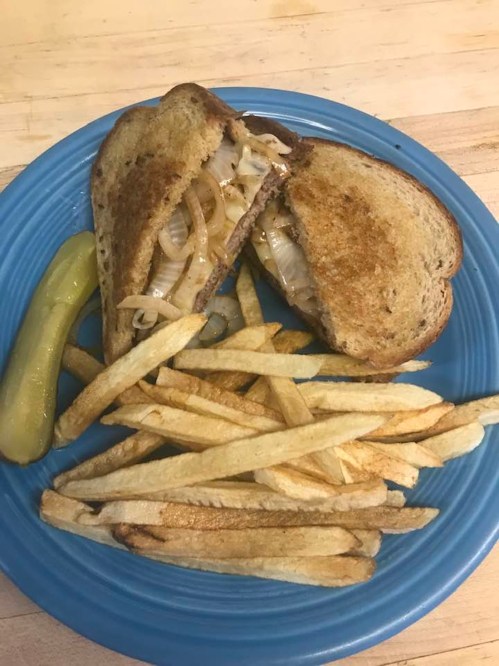 A toasted sandwich filled with onions, served with fries and a pickle on a blue plate.