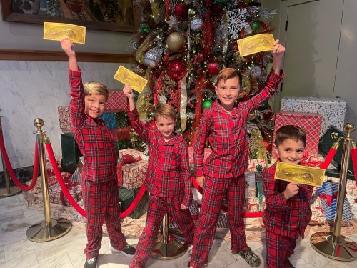 Four children in matching red plaid pajamas hold up golden tickets in front of a decorated Christmas tree and gifts.