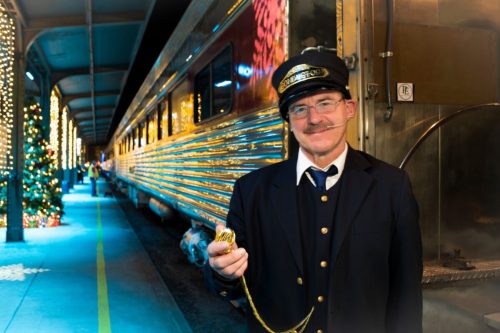 A train conductor in uniform holds a pocket watch, standing beside a beautifully lit train at a festive station.