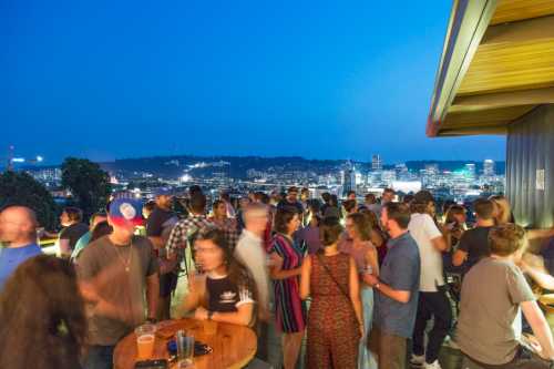 A lively rooftop gathering at night with people socializing and a city skyline in the background.