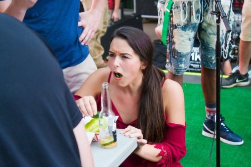 A woman in a red dress sits at a table, looking surprised while holding a drink, surrounded by a lively crowd.