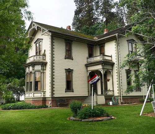 A large, historic two-story house with intricate woodwork, surrounded by greenery and a well-maintained lawn.