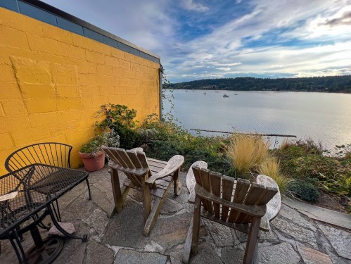 A cozy outdoor seating area with two wooden chairs and a table, overlooking a calm water view and a yellow wall.