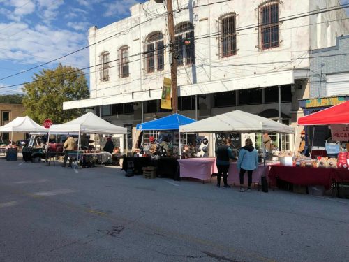 A bustling outdoor market with tents, vendors, and shoppers along a street in front of a historic building.