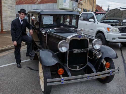 A man in a tuxedo stands next to a vintage black car on a street, with buildings in the background.