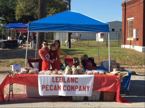 Two vendors at a market stand under a blue tent, showcasing pecan products with a sign that reads "Leblanc Pecan Company."