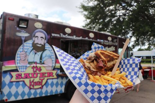 A hand holds a basket of fries and a sandwich in front of a food truck with a colorful logo.