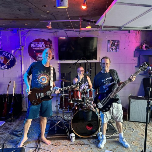 Three musicians in a garage setting: one on guitar, one on drums, and one on bass, with colorful lights and instruments around.