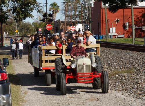A tractor pulls a wooden wagon filled with people along a gravel path near a train crossing.