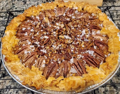 A freshly baked pecan pie with a golden crust, topped with pecans and a sprinkle of salt, cooling on a wire rack.