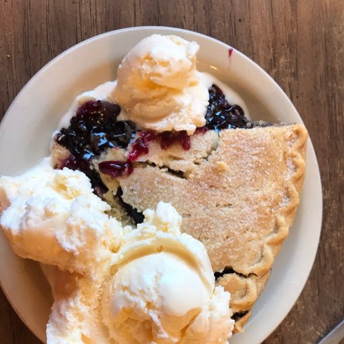 A slice of pie filled with dark berry filling, topped with two scoops of vanilla ice cream on a white plate.