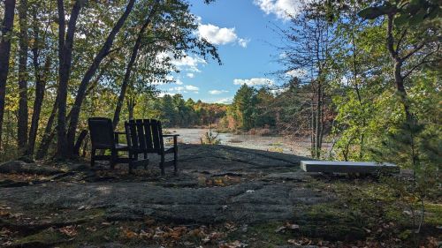 A serene outdoor scene featuring two chairs on a rocky ledge, surrounded by trees and a view of a calm, reflective water body.
