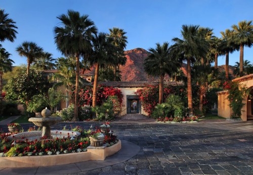 A scenic courtyard with palm trees, vibrant flowers, and a fountain, framed by a mountain backdrop under a clear blue sky.