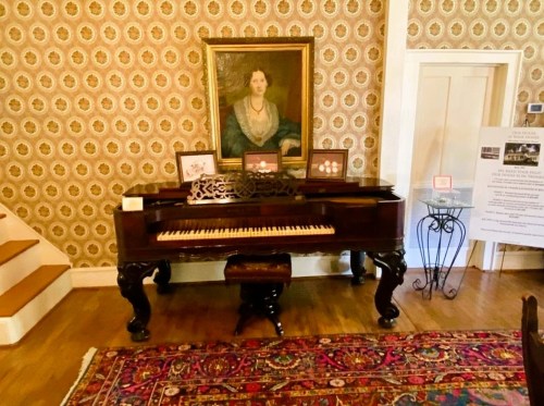 A vintage piano in a room with patterned wallpaper, a portrait above it, and a decorative rug on the floor.