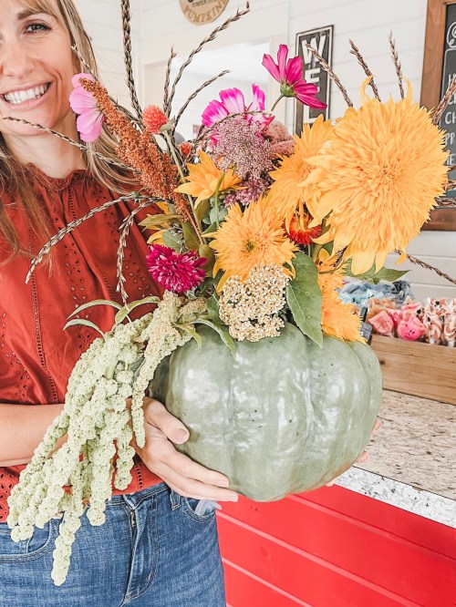 A person holds a large green pumpkin filled with vibrant flowers and greenery, smiling in a bright, cheerful setting.