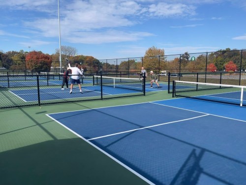 A sunny day at a tennis court with players engaged in a match, surrounded by colorful autumn trees.