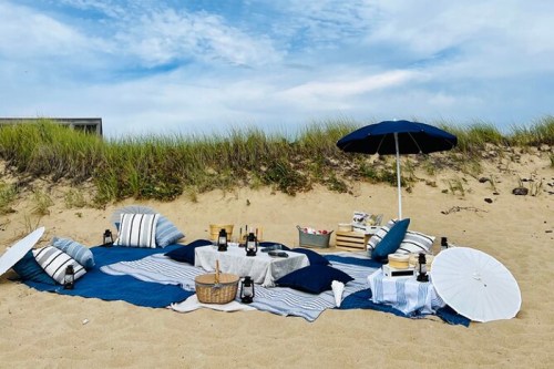 A cozy beach picnic setup with blankets, cushions, an umbrella, and picnic baskets on sandy ground.