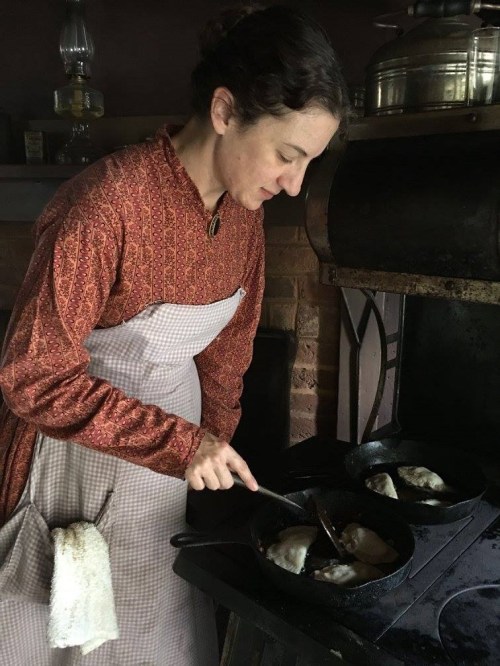 A woman in historical attire cooks over a stove, stirring food in cast iron pans.
