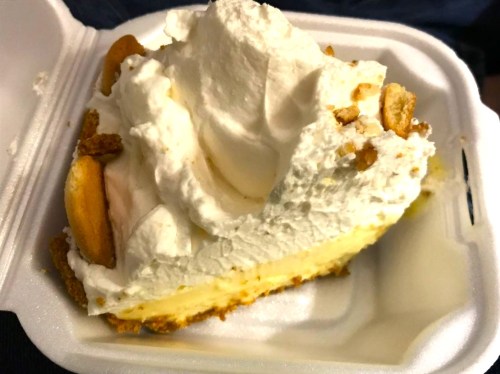 A slice of creamy dessert topped with whipped cream and crushed cookies in a takeout container.