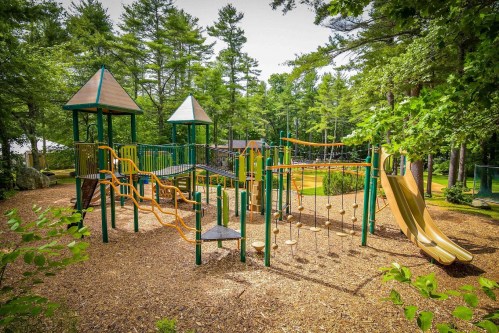A colorful playground with slides, climbing structures, and swings, surrounded by trees and mulch.