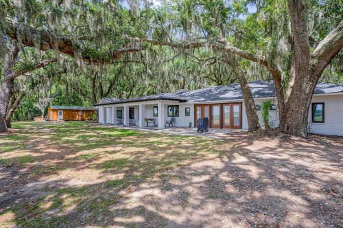 A spacious house surrounded by large trees with Spanish moss, featuring a patio and a separate shed in the background.