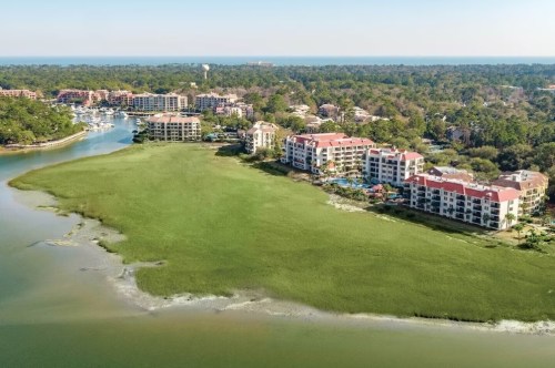 Aerial view of a coastal area with green marshland, buildings, and a marina under a clear blue sky.