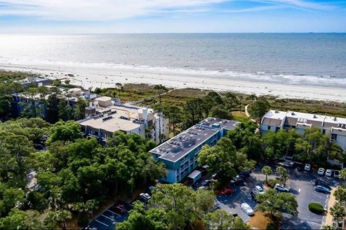 Aerial view of a beachside area with buildings, trees, and a sandy shore under a clear blue sky.