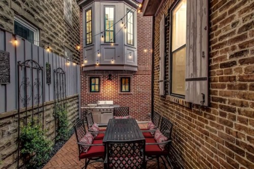 Cozy outdoor patio with a dining table, grill, and string lights, surrounded by brick walls and a unique upper balcony.