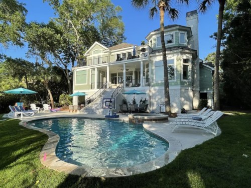 A spacious house with a pool, surrounded by palm trees and lounge chairs, set in a lush green backyard.