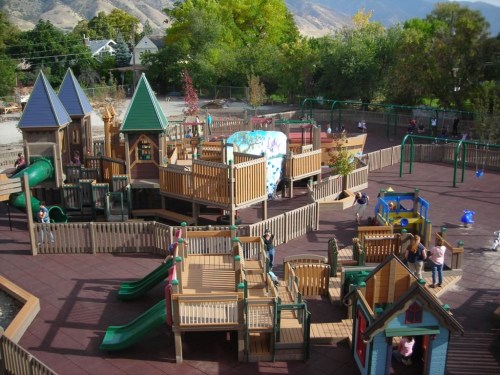 A colorful playground with various structures, slides, and climbing areas, surrounded by trees and mountains in the background.