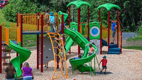 Children play on a colorful playground with slides, climbing structures, and adults supervising in a sunny outdoor setting.