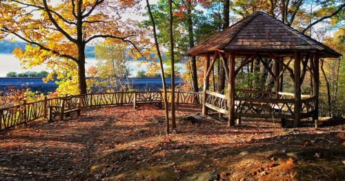 A wooden gazebo surrounded by colorful autumn trees and fallen leaves, overlooking a serene landscape.