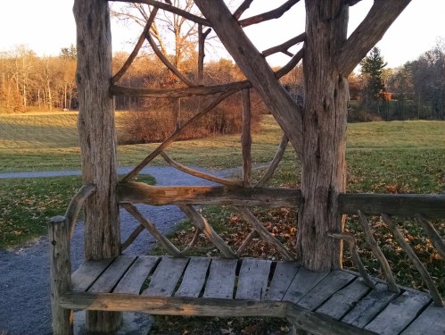 A wooden bench with a rustic design, surrounded by trees and a grassy field in a serene outdoor setting.