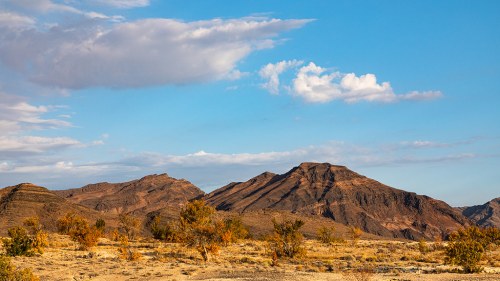 A scenic view of rugged mountains under a blue sky with scattered clouds, surrounded by arid landscape and sparse vegetation.
