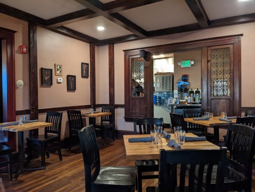 Cozy restaurant interior with wooden tables, black chairs, and framed artwork on the walls. Kitchen visible in the background.