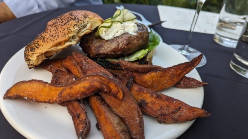 A plate with a poppy seed bun burger topped with lettuce and pickles, served with sweet potato fries.