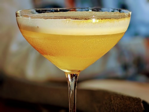 A close-up of a golden cocktail in a elegant glass, with bubbles and a frothy top, set against a blurred background.