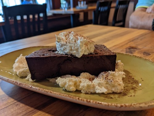 A slice of rich chocolate cake topped with whipped cream, served on a green plate with dollops of cream on the side.