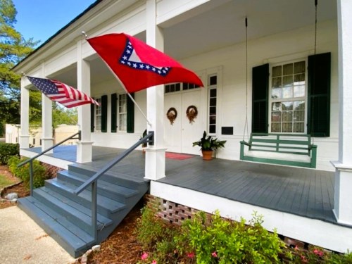 A porch with two flags (American and Arkansas), a swing, and potted plants, featuring white walls and green shutters.
