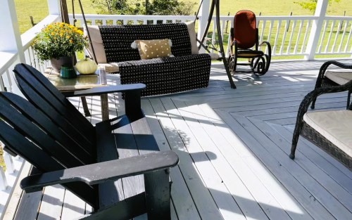 A cozy porch with a black Adirondack chair, a wicker sofa, and a rocking chair, surrounded by greenery and flowers.
