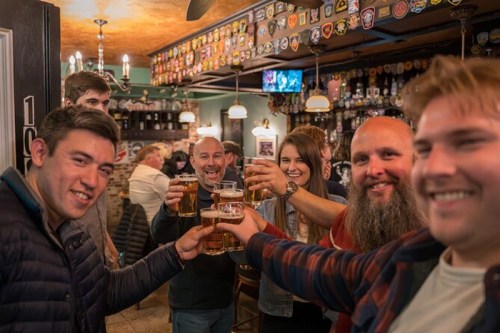 A group of friends cheerfully toasting with beers in a lively pub filled with memorabilia.