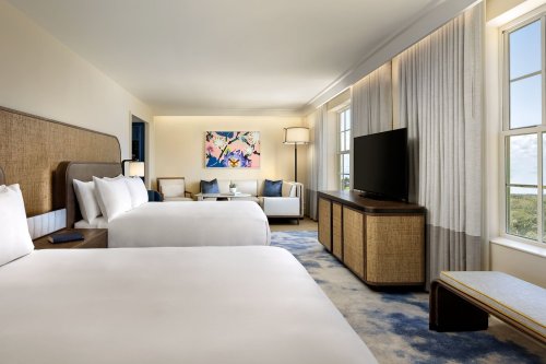 A modern hotel room featuring two beds, a seating area, large windows, and colorful artwork on the wall.